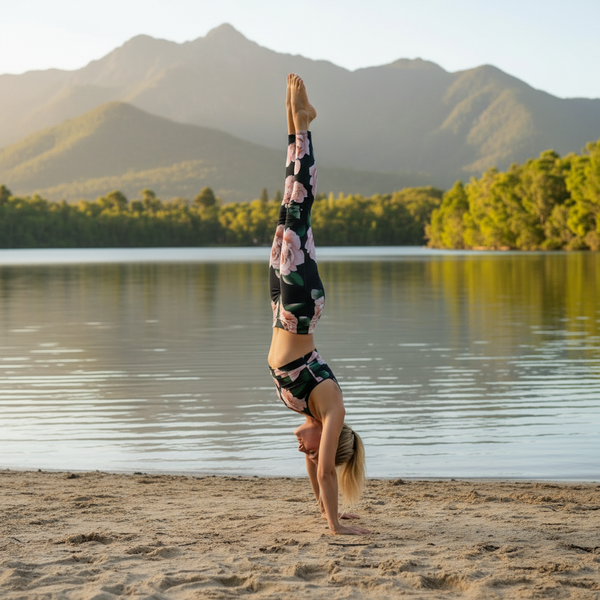 A woman performs a handstand yoga pose on a sandy beach with a calm lake and green mountains in the background. She is wearing a matching floral print activewear set.